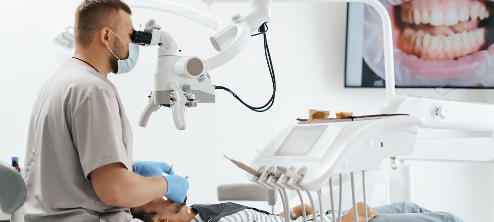 young-man-with-patient-bib-dental-chair-dentist-who-sits-him-he-looks-his-teeth-using-dental-microscope-holds-dental-bur-mirror
